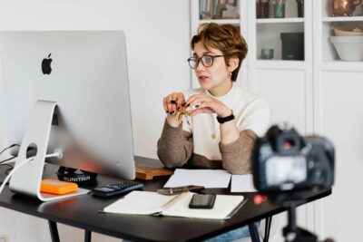 CRM for Photographers: What to Look For 97 CRM for photographers A professional woman engaged in a virtual meeting setup at her home desk, using a computer and camera.