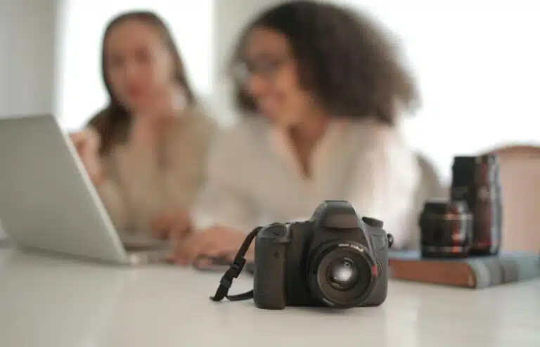 Go High Level vs Dubsado Close-up of DSLR camera with two women collaborating on a laptop indoors.