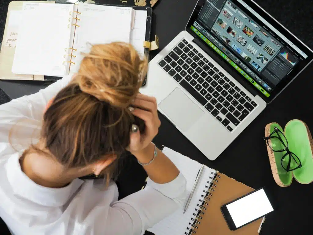 small business CRM Overhead view of a stressed woman working at a desk with a laptop, phone, and notebooks.